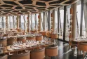 The 7 panoramic restaurant at La Cité du Vin in Bordeaux featuring elegant light wood tables, caramel-colored chairs, wine glasses, modern geometric ceiling, and floor-to-ceiling windows overlooking the city