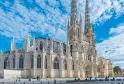 Bordeaux Cathedral of Saint-André, a white Gothic structure featuring two main towers, ornate facade with stained glass windows and rose windows, surrounded by visitors in a paved courtyard under a cloudy blue sky.