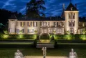Château Bellefont Belcier in Bordeaux lit up at night, featuring a pale stone facade with pointed towers, central access stairs and illuminated fountains in the foreground