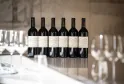 Six bottles of red wine from Château de Ferrand vintage years 2012 to 2017 lined up on a white table with blurred wine glasses in the background, in an elegant Bordeaux setting.