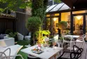 Elegant terrace of a boutique hotel in Bordeaux featuring white dining tables, modern chairs, landscaped garden, ambient lighting, and illuminated glass doors opening to the restaurant-bar interior.