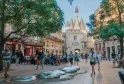 Cailhau Gate, a historic monument in Bordeaux, seen from a shaded public square with visitors, cafés, and ground sculptures