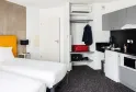 Modern studio apartment at a hotel in Talence, Bordeaux, featuring a white bed with yellow headboard, equipped kitchenette, storage shelves, and air conditioning in minimalist contemporary design