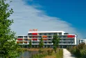Mer & Golf City Bordeaux Bruges apartment hotel, modern white and red building with balconies overlooking a canal, surrounded by greenery under blue sky.