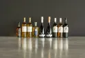 Nine bottles of Bordeaux wine arranged in a row on a gray surface, featuring golden white wines, red wines, and rosés from Château d'Arche and other Bordeaux châteaux.