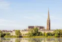 Panoramic view of the Basilica of Saint-Michel in Bordeaux with its Gothic spire towering over the urban landscape, surrounded by Haussmann-style buildings and reflected in the Garonne river waters.