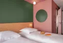 Modern hotel room at Eklo Bordeaux featuring two twin beds with natural wood headboard, color-blocked walls in soft green and pink, round green mirror, and minimalist design.