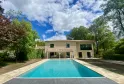Modern villa with rectangular swimming pool surrounded by lawn and trees, beige facade with covered terrace, cloudy blue sky.