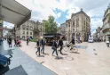 Busy square in Bordeaux with pedestrians, cyclists and shops, lined with Haussmann-style buildings under a cloudy sky.