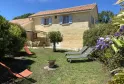 Rear view of a beige stone house with tiled roof, surrounded by a landscaped garden featuring lounge chairs, pergola, and lush vegetation, under clear blue sky.