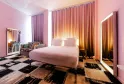 Modern bedroom at Mama Shelter Bordeaux featuring a white bed, soft pink walls, gradient orange and gold curtains, illuminated mirrors, striped black and white armchair, and geometric black and beige carpet