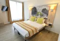 Modern hotel room in Bordeaux featuring a double bed with white linens and green cushions, large artwork of a castle above the headboard, striped curtains on the window, wall-mounted flat screen TV, and light wooden flooring.