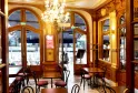 Elegant interior of QuatrèS Café in Bordeaux featuring golden wood paneling, crystal chandelier, wooden glass doors, and classic furniture with outdoor terrace visible.