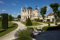 White Belle Époque mansion with black domes surrounded by manicured gardens, lounge chairs in foreground and shaded terrace in background