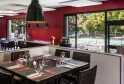 Modern restaurant featuring vibrant red walls, elegantly set black dining tables with glassware and place settings, dark pendant light fixture, and floor-to-ceiling windows overlooking a swimming pool and outdoor lounge area.