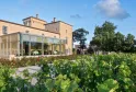 Exterior view of the Lalique restaurant at Château Lafaurie Peyraguey in Bordeaux, featuring a modern glass extension overlooking vineyards, with historic stone buildings and blue sky