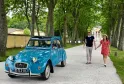 A turquoise Citroën 2CV with an open roof parked under a tree-lined avenue in Bordeaux, with a couple walking nearby and historic buildings in the background.