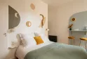 Modern bedroom at La Maison Fernand in Bordeaux featuring a white bed, minimalist décor, round gold mirrors, mustard yellow and sage green accents, wall shelf and wooden stools.