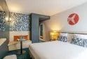 Modern hotel room at Ibis Styles Bordeaux Lac Bruges featuring a white bed, orange bench seating, blue and white geometric patterns, and minimalist coordinated décor.