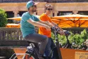 Two people riding an orange Holland Bikes rental bicycle in Bordeaux, with orange umbrellas and flowers in the background