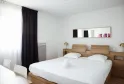 Modern hotel room in Bordeaux featuring a double bed with white linens, natural wood headboard, wall mirror, black and white curtains, gold bedside lamps, and minimalist décor.
