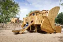 Wooden boat-shaped playground with slide, children playing, surrounded by wooden fence, at Forêt du Bourgailh in Bordeaux.