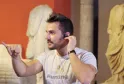 A man in a beige Hunting t-shirt talks on the phone in a museum exhibition, surrounded by ancient sculptures and orange walls.