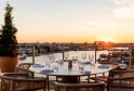 Rooftop terrace of a hotel with a round table set for dining, wicker chairs, glassware and place settings, overlooking Bordeaux at sunset with the Garonne river in the background