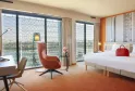 Spacious suite at Radisson Blu Hotel Bordeaux featuring two white beds, coral red armchair, floor-to-ceiling windows overlooking the city, and modern minimalist décor.