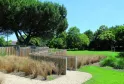 Public park along the Jalle de Gajac waterfront in Bordeaux featuring green lawns, ornamental grasses, wooden and metal fencing, mature trees and clear sky