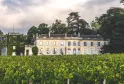 View of Château du Taillan near Bordeaux, classical stone façade overlooking vineyards in the foreground, surrounded by lush greenery and century-old trees.
