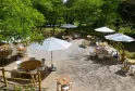 Shaded outdoor terrace at Château Léognan featuring wooden tables, white chairs and white umbrellas, surrounded by lush green trees.