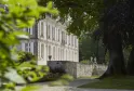 View of Château de La Dauphine in Bordeaux featuring its white neoclassical facade with columns, surrounded by lush greenery and ancient trees, accessed by stone steps.