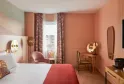 Elegant hotel bedroom with coral walls, white bed with gold palm-shaped headboard, pink armchair, floral curtains, wooden desk and wall-mounted television.