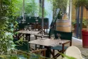 Shaded outdoor dining terrace at Les Tables Vatel restaurant in Bordeaux featuring wooden tables, beige umbrellas, lush greenery, and a wine barrel in the background
