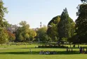 View of Bordeaux Public Garden featuring lush green lawns, tall trees, relaxing visitors, a monument with statue in the background and historic buildings under clear sky.
