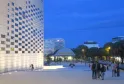 Night view of La Méca in Bordeaux featuring its illuminated white checkered facade, a paved public plaza with visitors, and modern buildings against a twilight sky.