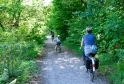 Cyclists riding on a shaded forest trail near Bordeaux, in the Bois du Déhès and Moulin du Moulinat area, surrounded by lush vegetation.