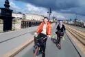 Two smiling cyclists riding bikes on a dedicated bike lane crossing a historic bridge in Bordeaux, with classical buildings and a cathedral in the background