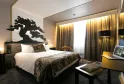 Modern hotel room featuring a queen-size bed, black dragon wall art, golden curtains, desk with laptop, and contemporary black and gold furnishings.