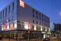 Modern facade of Ibis Bordeaux Centre Bastide hotel at night, featuring contemporary gray and white architecture, illuminated red signage, and parked cars in front