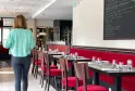 Interior of a Bordeaux restaurant showing a woman in a turquoise shirt standing before a row of tables with red banquettes, wooden chairs, water glasses, and a chalk menu board on the wall.