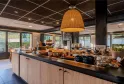 Breakfast buffet in the Campanile Bordeaux Le Lac restaurant dining area, featuring pastries, breads, and various food items on a long counter with wicker pendant light, large windows overlooking nature, and light wood furnishings.