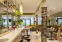 Modern restaurant interior in Bordeaux featuring a light wood bar, decorative floral columns, green plants, olive-green seating, and large windows overlooking the garden.