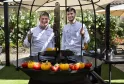 Two chefs in white uniforms pose in front of a circular brazier filled with colorful tomatoes in a flowering garden with pergola in Bordeaux.