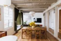Bright loft interior with exposed wooden beams, wooden dining table with rattan chairs, large windows, open kitchen, and minimalist décor in Bordeaux.