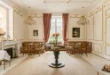 Elegant neoclassical drawing room at Château Léognan in Bordeaux, featuring cream walls, gilt wood paneling, two ornate chandeliers, red curtains, round central table, fireplace, antique portraits, and wicker furniture.